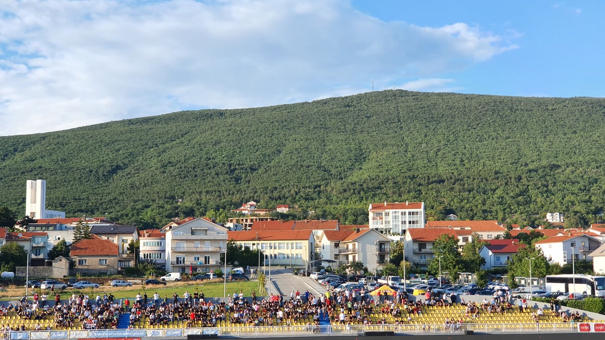 Crowded football stadium in Posušje with lush hills and townscape in the background.